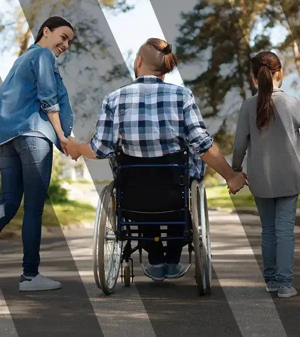 A man in a wheelchair having a nice stroll outside while holding hands with his wife and daughter .