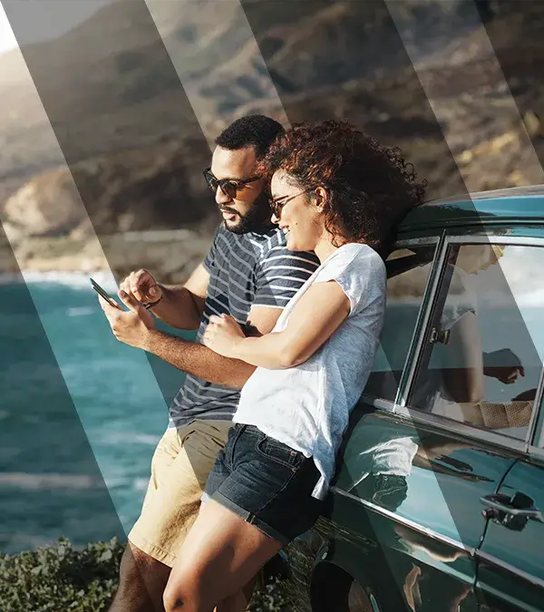 A couple looking at mobile phone leaning back on a car while at the beach.