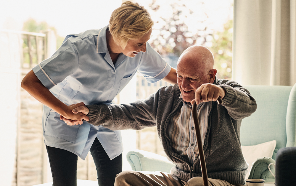 Female nurse helping an elderly man get up off of a chair.